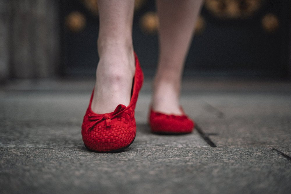 Nahaufnahme einer Person, die in TWIST Red-Schuhen von ZAQQ auf einem Steinpflaster läuft. Dabei werden das Velourleder-Finish und die zarten Schleifen hervorgehoben. Das lebhafte Schuhwerk steht im Mittelpunkt, während der Hintergrund sanft verschwommen bleibt.