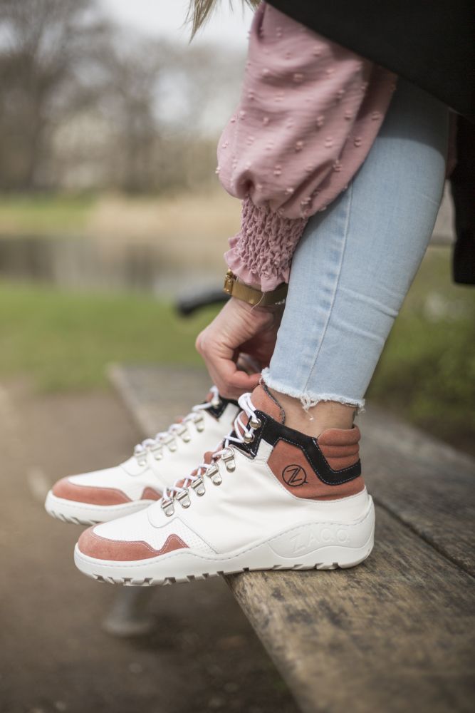 Eine Person sitzt im Freien auf einer Bank. Sie trägt hellblaue Jeans und ein rosa strukturiertes Oberteil und schnürt die VAQATION Rosa Waterproof Sneaker von ZAQQ in Weiß und Braun. Im Hintergrund ist eine verschwommene Parklandschaft mit Bäumen und Gras zu sehen, die einen friedlichen Moment einfängt.